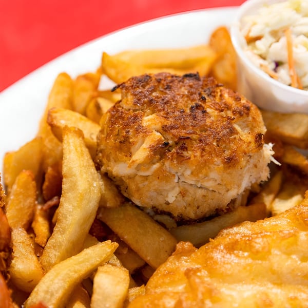 Go Fish! crab cake, golden fries, and coleslaw on a white plate with a red background.