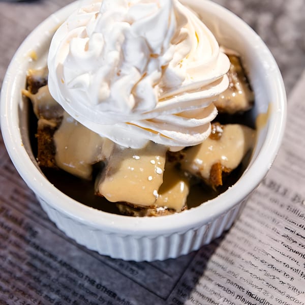 Sticky toffee pudding dessert with whipped cream in a white ramekin on a newspaper-patterned surface.