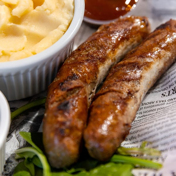 Two grilled sausages with mashed potatoes and a side of sauce, served on newspaper.