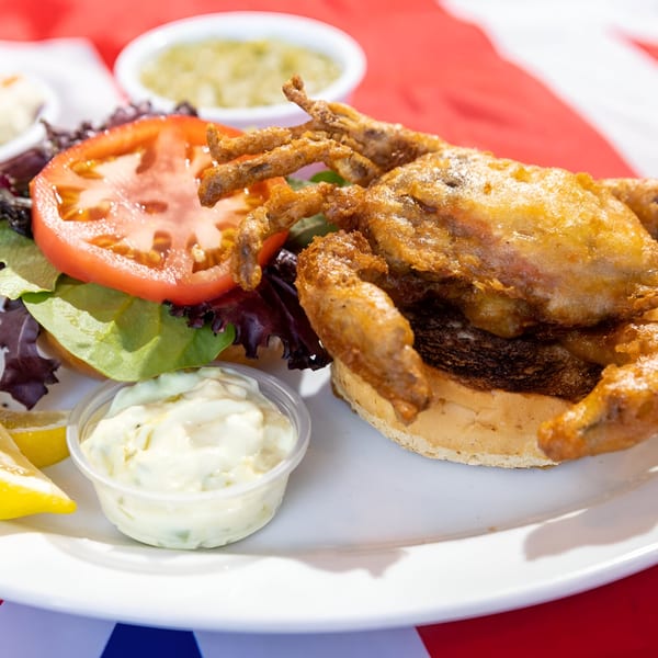 A fried soft-shell crab sandwich with lettuce, tomato, and tartar sauce on a white plate.